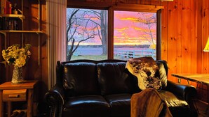 Et hyggeligt interiør i en hytte med en lædersofa, en træreol og et stort vindue med udsigt over en solnedgang over søen.