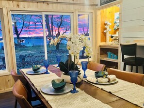 En matplats med utsikt över en solnedgång över en sjö, ett bord dukat med blått glas, vita orkidéer och en kaffestation.