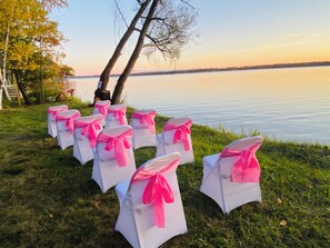 Rows of white chairs with pink sashes arranged on a grassy area by a lake. - Cozy Waterfront 4-bedroom Cottage. Best Sunsets! 10 mins South of Mackinaw City! (Carp Lake)