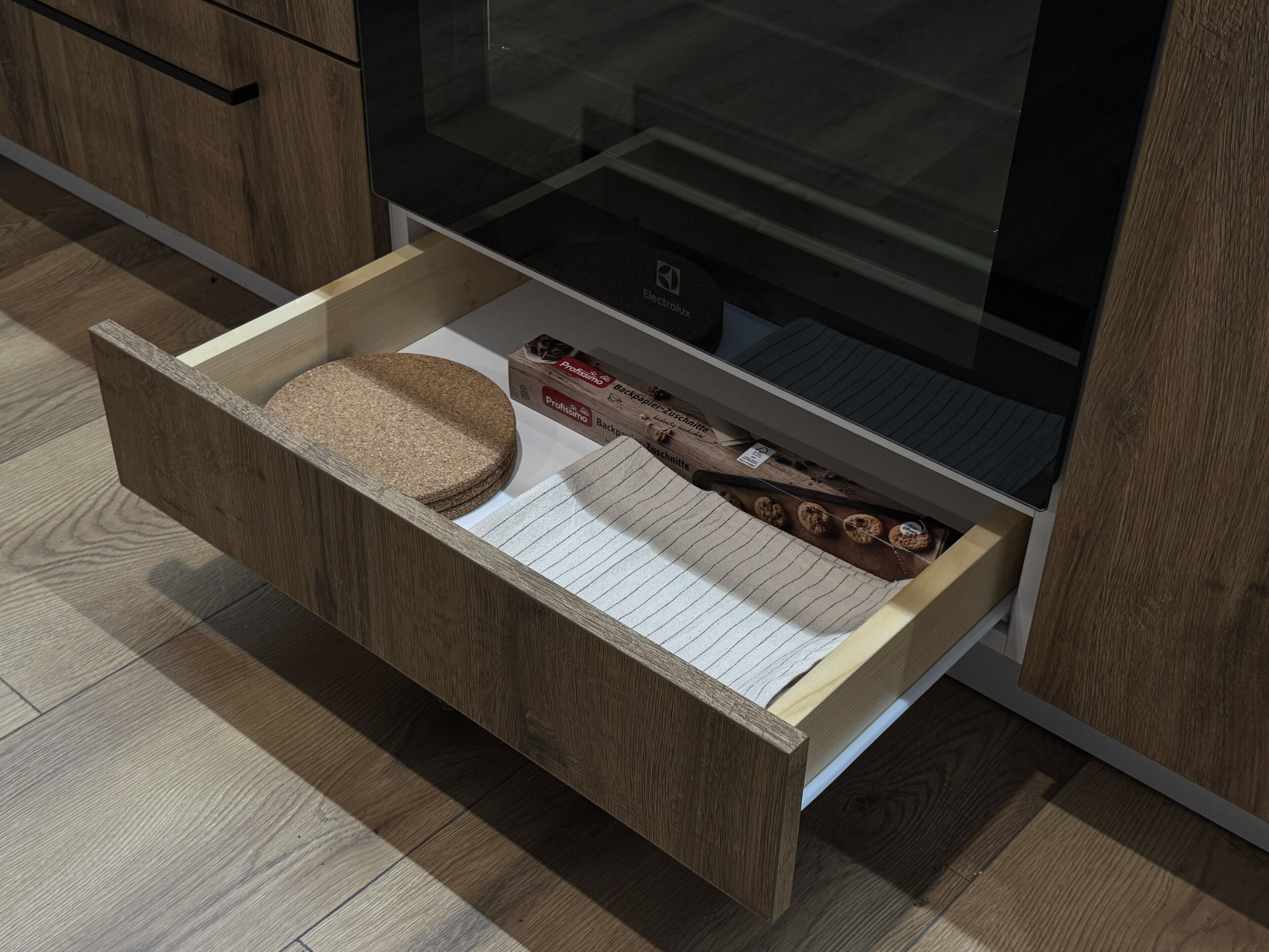 A wooden drawer with a cork mat, lined with white paper, containing a box labeled "Profissimo Backpulver-Zuschne" and some cookies.