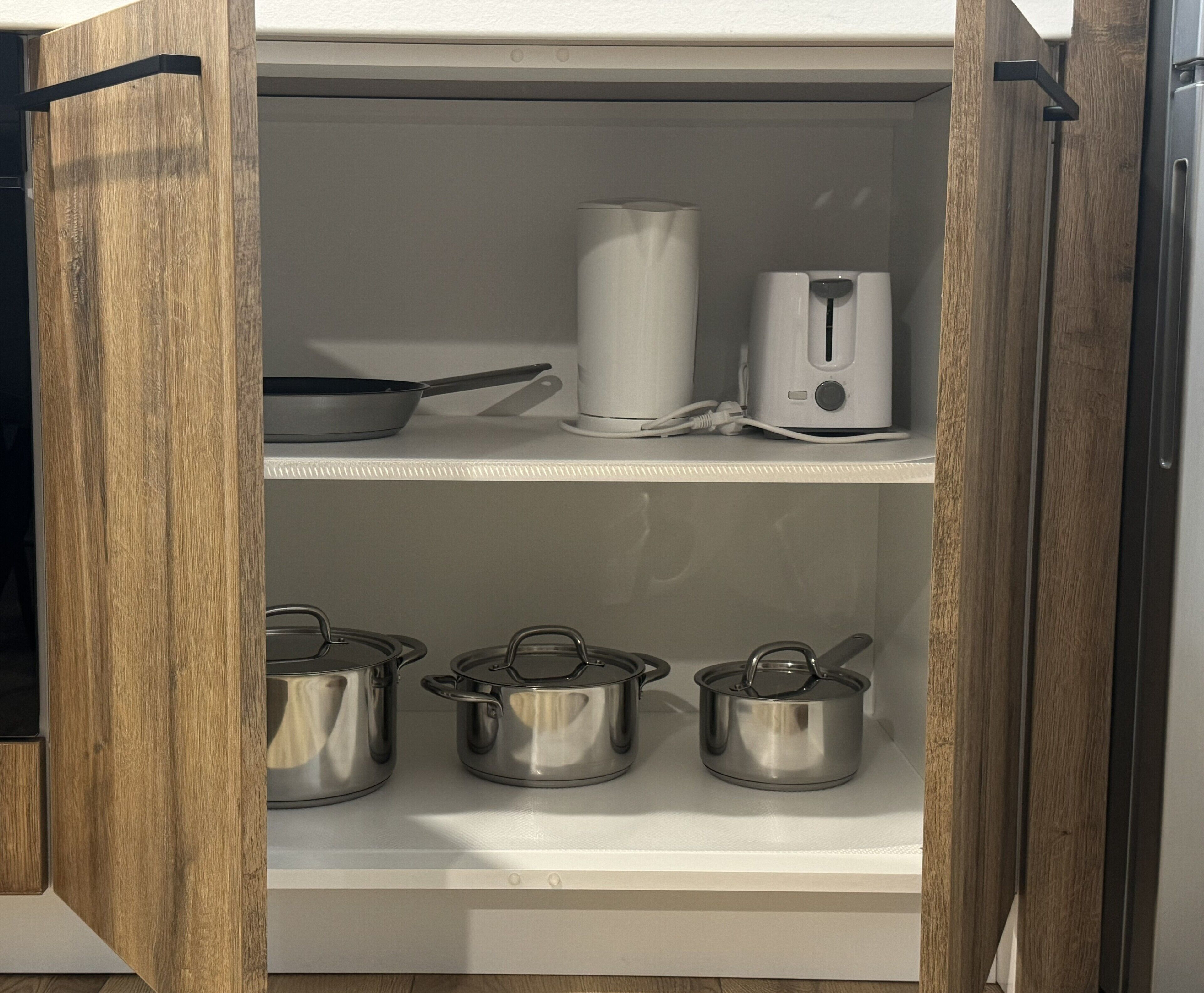 Un armario de cocina con tres ollas de acero inoxidable en el estante inferior y una tetera eléctrica y una sartén blanca en el estante superior.