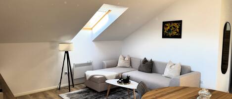 A modern loft apartment with a sloped ceiling, a skylight, a gray sofa, a wooden dining table, and a black pendant light.