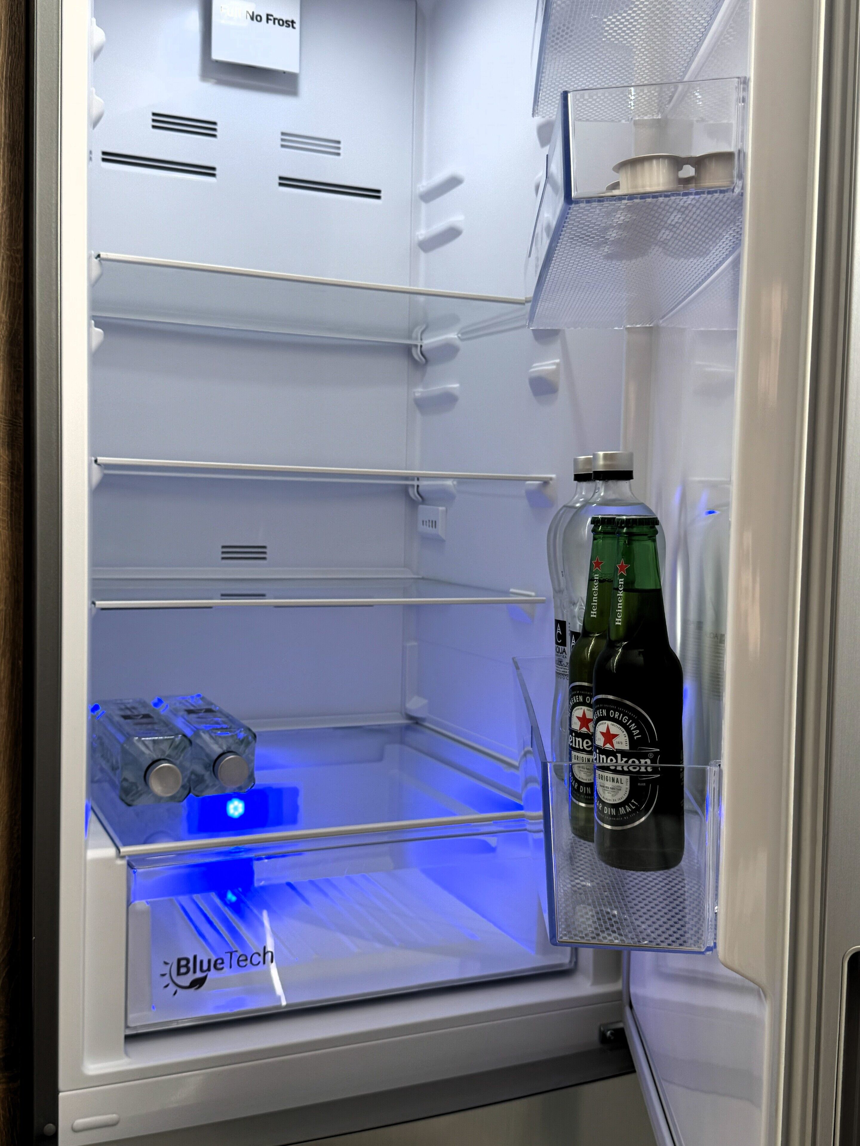 Un réfrigérateur vide éclairé d’une lumière bleue, une bouteille de Heineken et le logo BlueTech.
