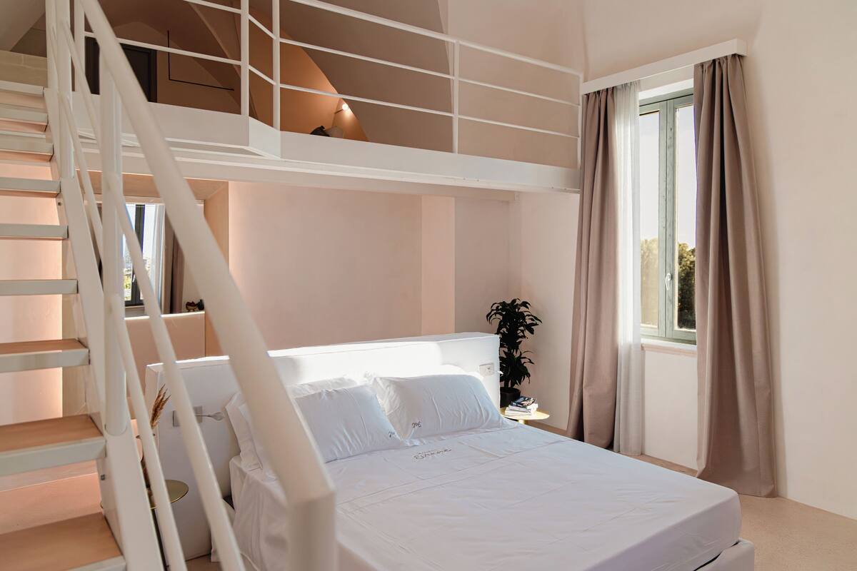 A bedroom with a bunk bed, a staircase, a bedside table with a lamp, and a window with curtains.