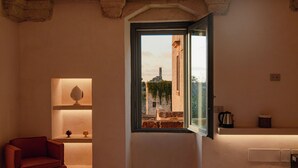 A bedroom with a bed, a chair, a window with a view of buildings, and a shelf with a lamp and a container. - Masseria Eirene (Lecce)