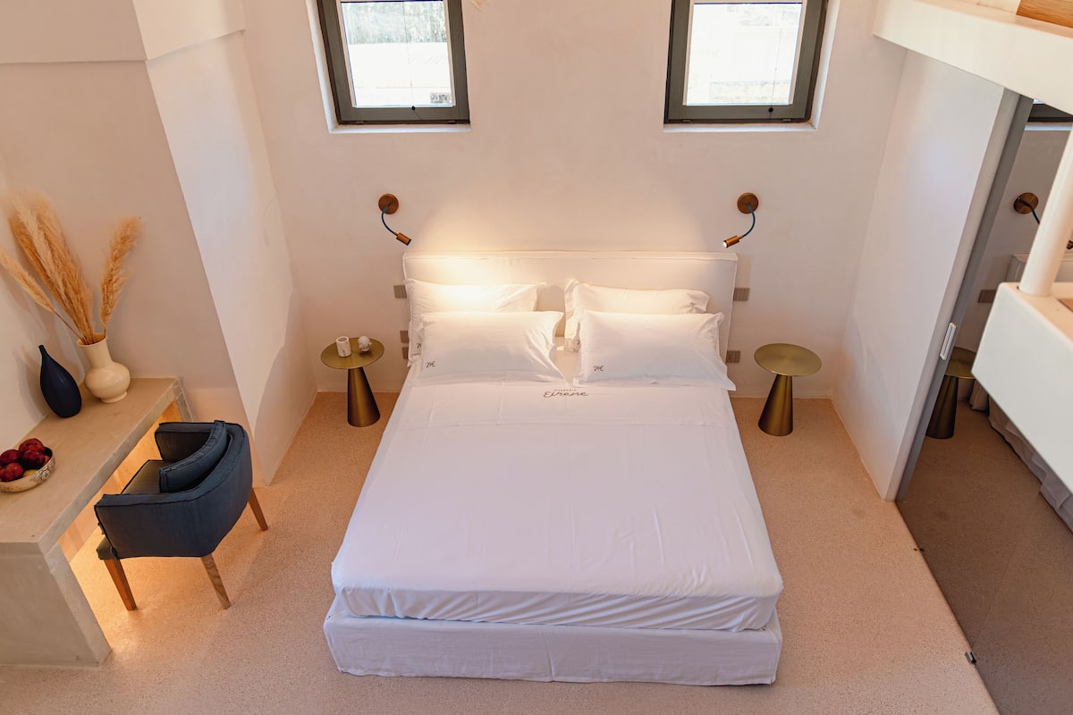 A neatly made bed with white linens, a bedside table with a vase and a bowl of fruit, and a chair.