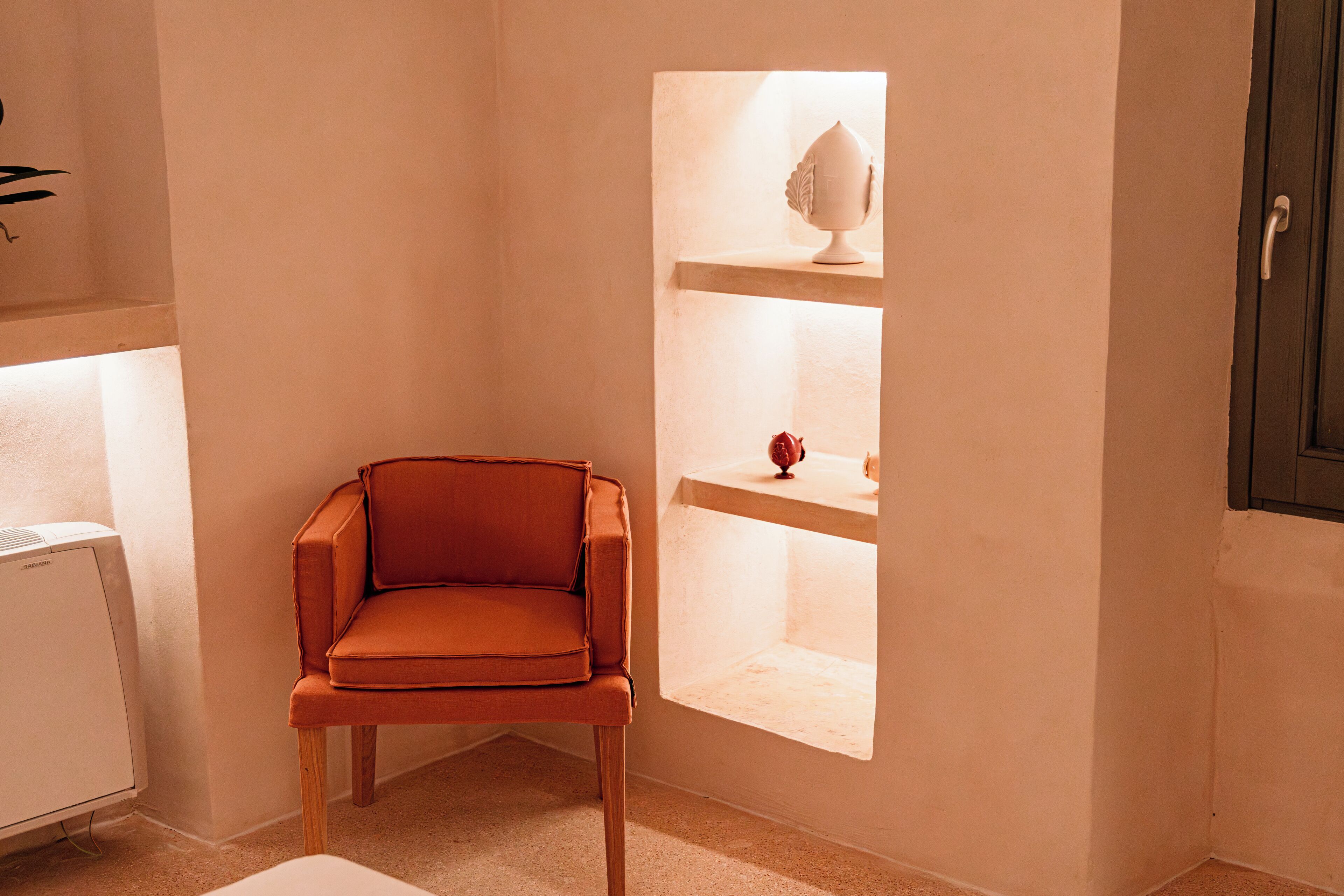 A corner room with a single orange chair, a small shelf with decorative items, and a white air conditioning unit.