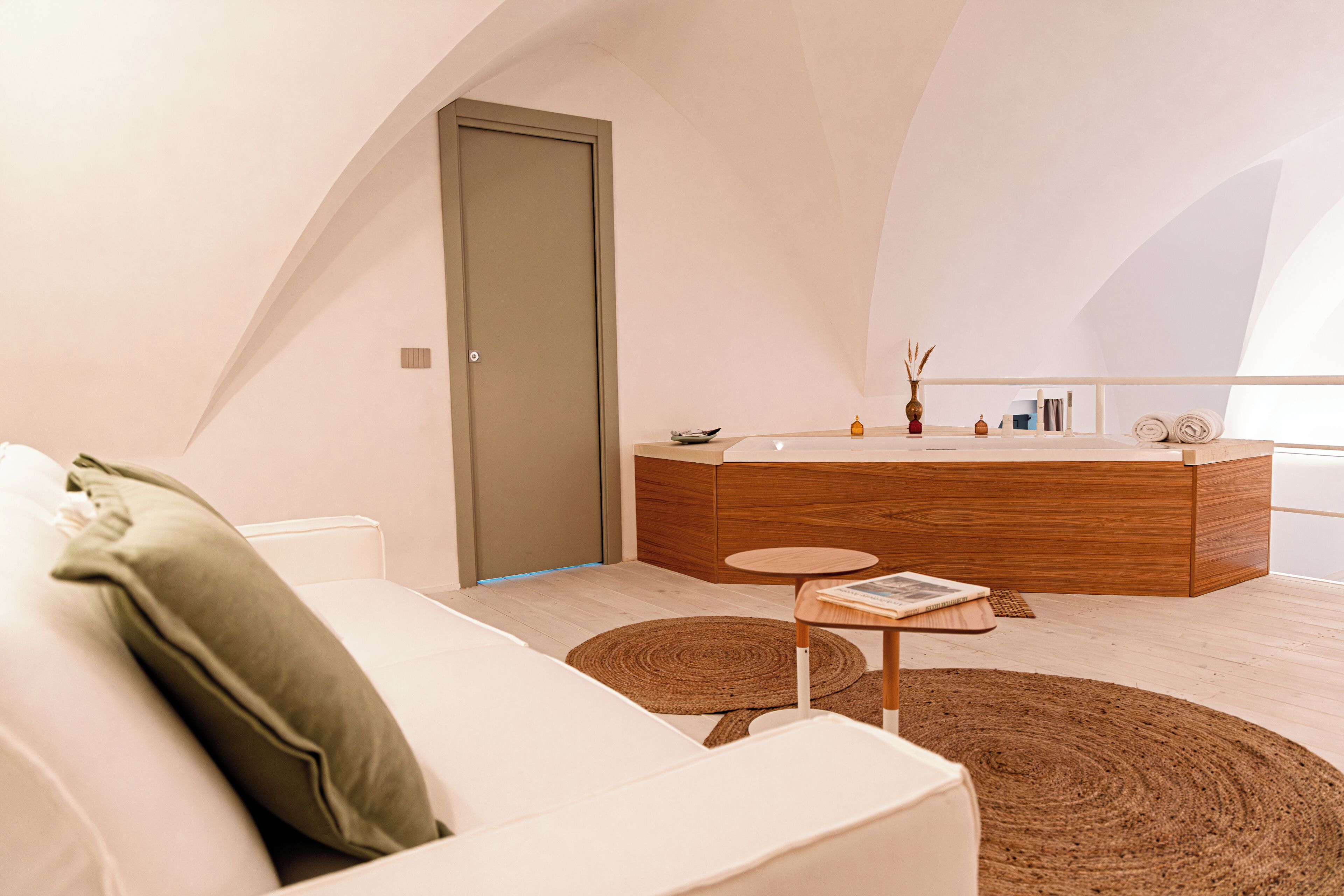 A modern bedroom with a white sofa, a wooden bedside table, a round rug, and a large window.