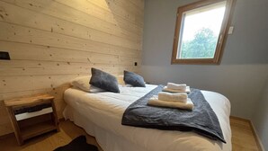 A neatly made bed with white linens, a dark striped blanket, and a window with a view of trees. - Magnificent Chalet - Swimming pool & exceptional view (Les Avanchers-Valmorel)