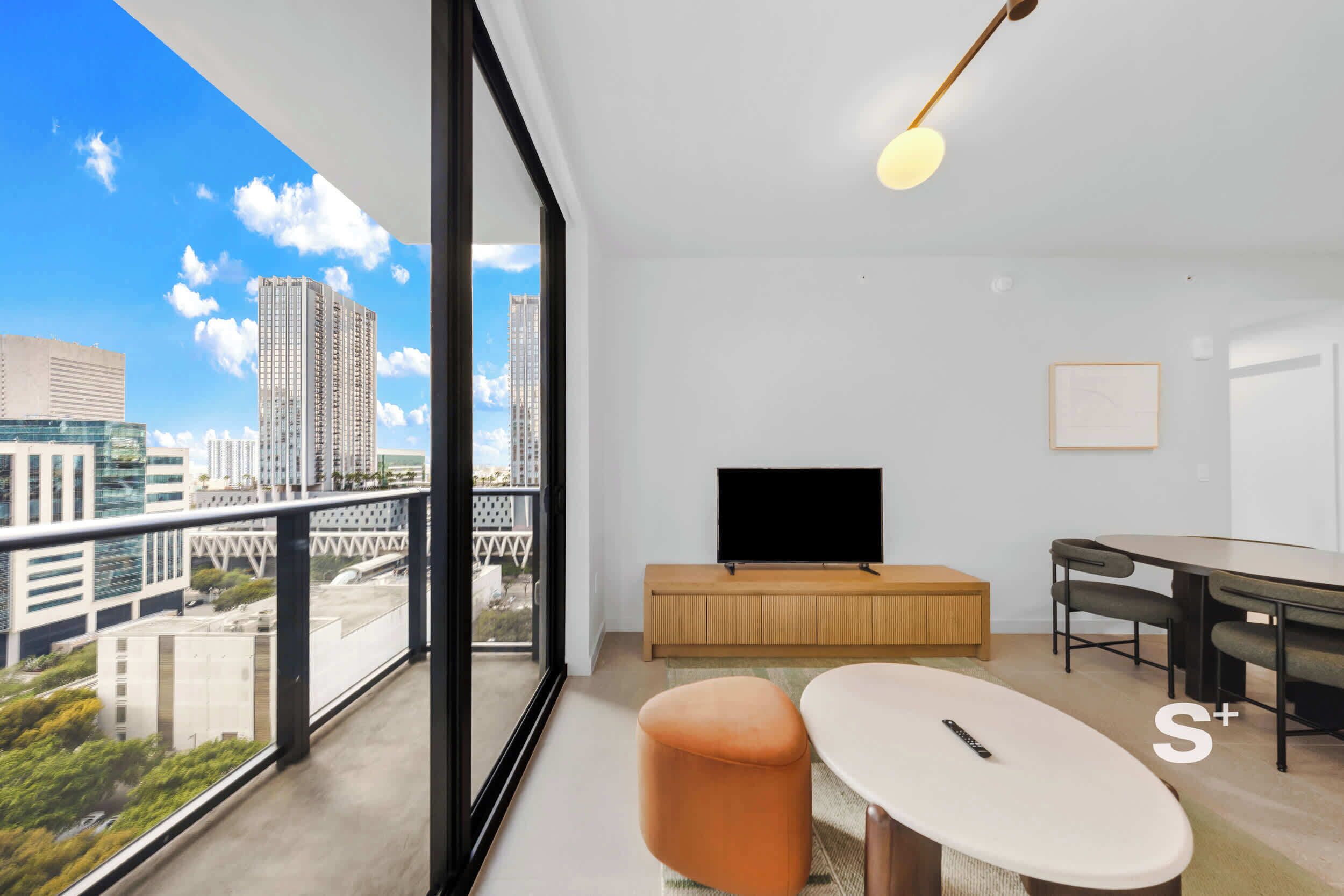 A modern living room with a large window, a flat-screen TV, a dining table, and a sofa.
