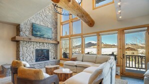 A cozy living room with a stone fireplace, a large window with a mountain view, and a wooden ceiling beam. - Walk to Lift! Luxe, Updated CBMR Home w/ Mtn Views (Gunnison)