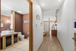 Ein Hotelgang mit Holzboden, ein Badezimmer mit weißem Waschbecken und WC, und ein Bett im Hintergrund.