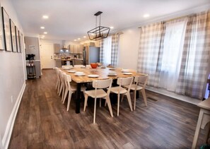 Uma área de refeições com uma mesa e cadeiras de madeira, uma cozinha ao fundo e uma janela com cortinas.