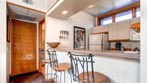 A compact kitchen with a wooden door, a microwave, a refrigerator, and a coffee maker. There are three metal bar stools with wooden seats.