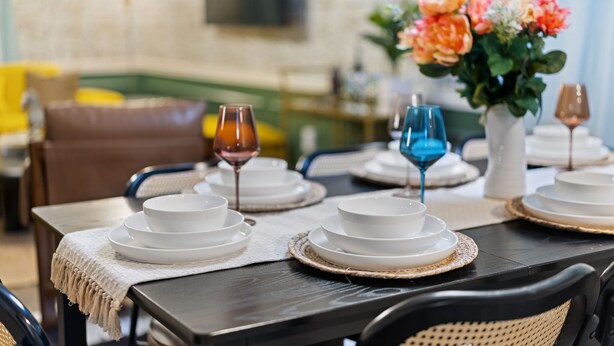 Una mesa de comedor dispuesta con platos blancos y cristalería azul, rodeada de sillas de mimbre.