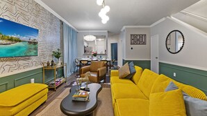 Una sala de estar con un sofá amarillo, una mesa de centro con libros, un televisor que muestra una escena tropical y una zona de comedor con un candelabro.