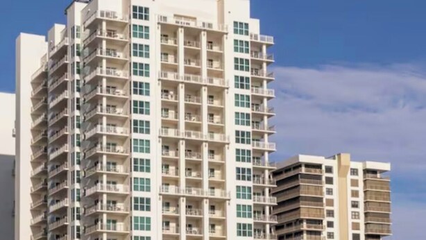 Un hôtel à plusieurs étages doté de balcons, une plage avec des chaises longues et des parasols, et des palmiers.