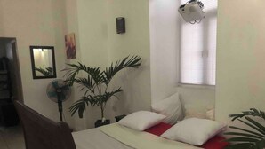 Ein Schlafzimmer mit einem Bett, einem Standventilator, einer Topfpflanze, einem Fenster mit Jalousien und einem an der Wand montierten Projektor.