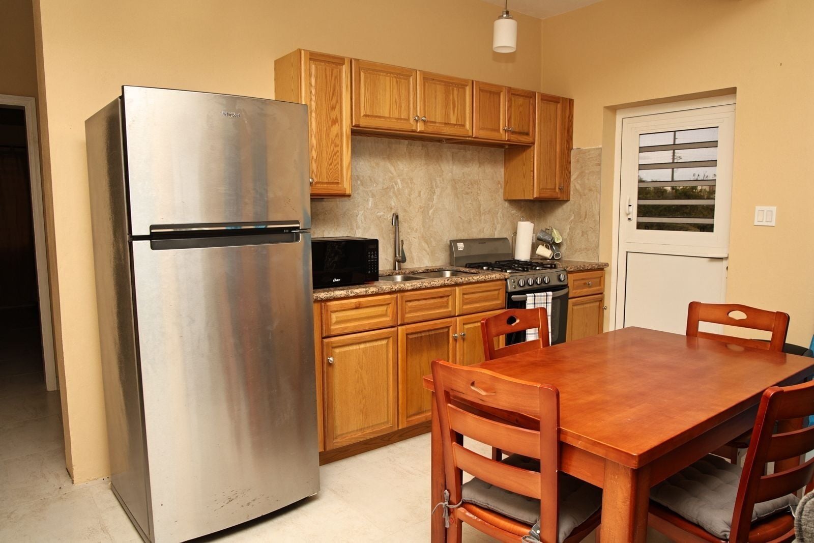 A compact kitchen with wooden cabinets, a stainless steel refrigerator, a microwave, a stove, and a dining table with chairs.