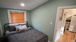 A bedroom with a bed, a window with blinds, a bedside table, and a door leading to a kitchen area. - Contemporary Countryside Home, Minutes to Concord (Loudon)