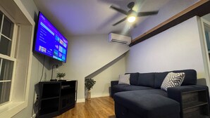 A modern living room with a flat-screen TV mounted on the wall, a ceiling fan, and a dark-colored sofa with decorative pillows. - Contemporary Countryside Home, Minutes to Concord (Loudon)