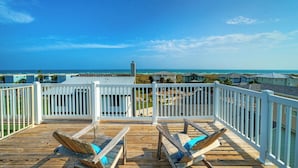 Eine Holzterrasse mit zwei Liegestühlen, einem weißen Geländer und Blick auf das Meer und Strandhäuser.