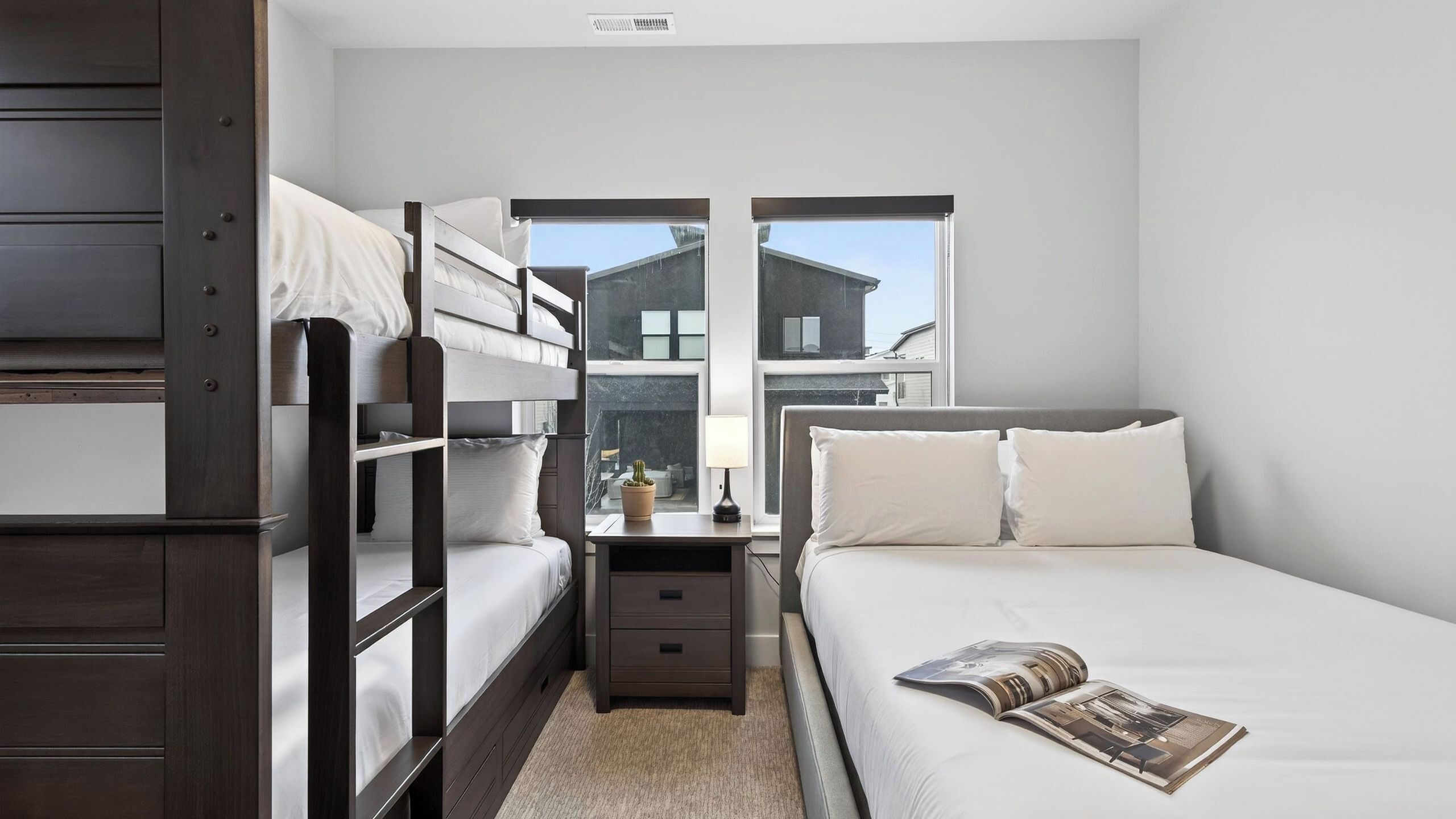 A room with bunk beds, a bedside table with a lamp, and a window with a view of buildings.