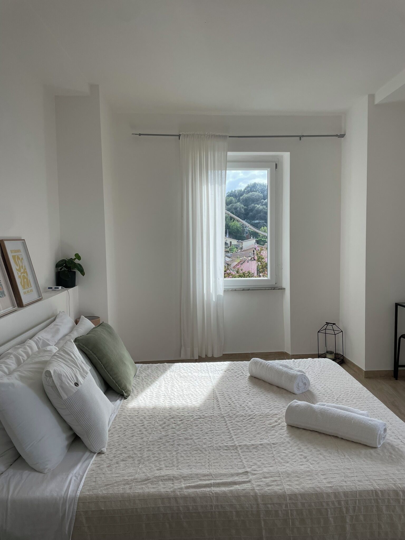 A bedroom with a large bed, white curtains, a view of a landscape, and a small table with a lamp.