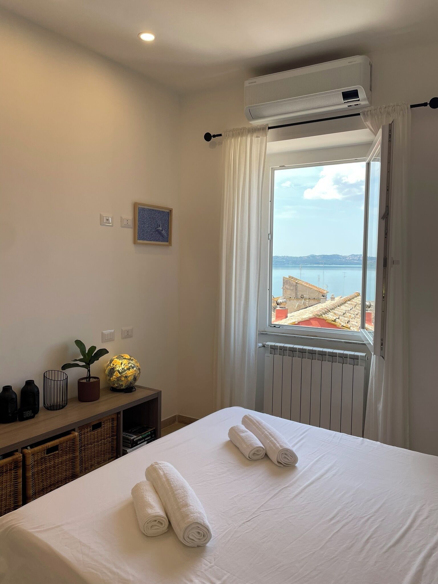 A hotel room with a bed, a window with a view of the sea, an air conditioning unit, and a small table with a plant and a decorative item.