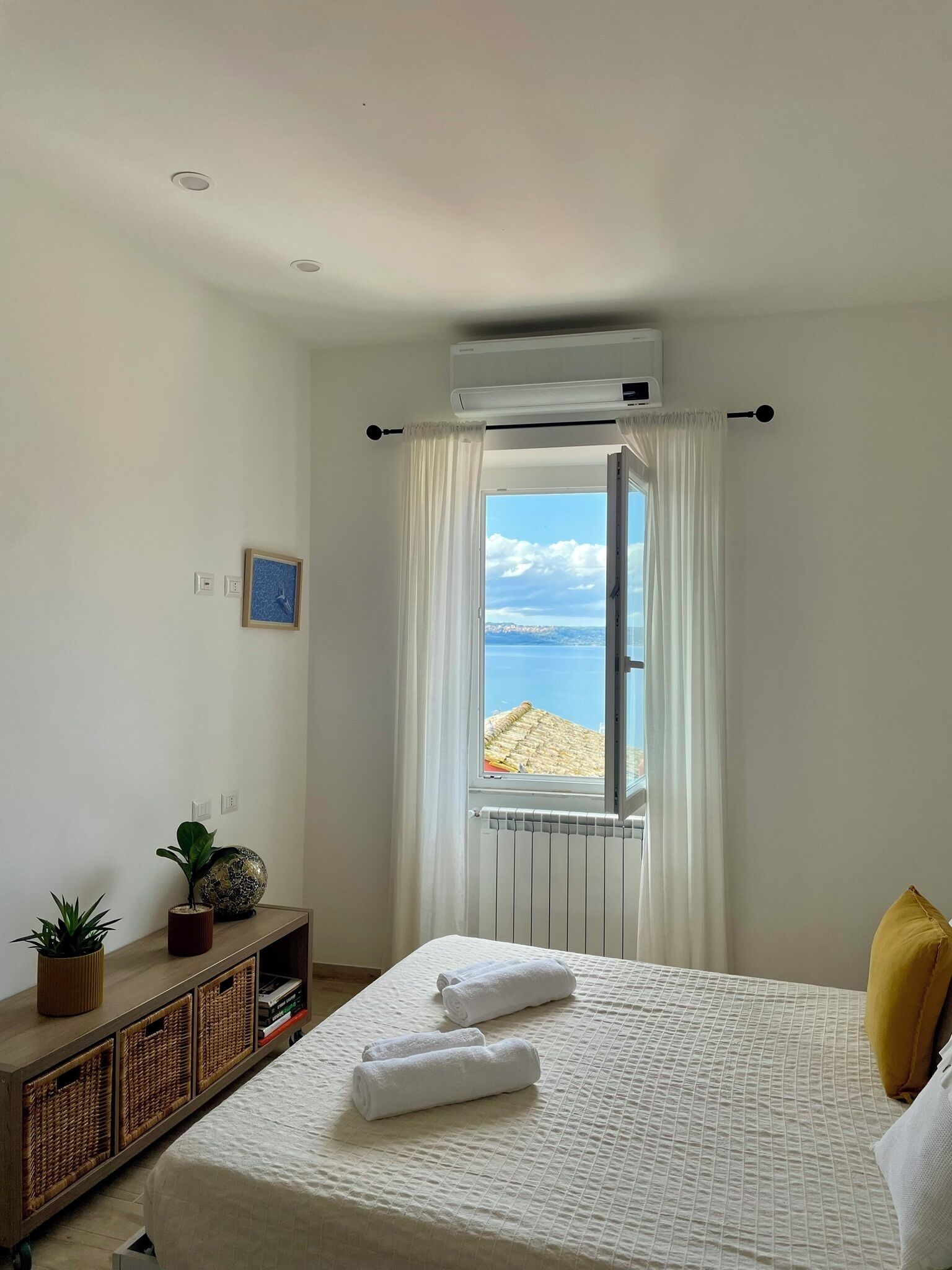 A bedroom with a bed, a window with a view of the beach, a radiator, and a small plant on the sideboard.