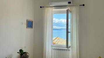 A bedroom with a bed, a window with a view of the beach, a radiator, and a small plant on the sideboard.