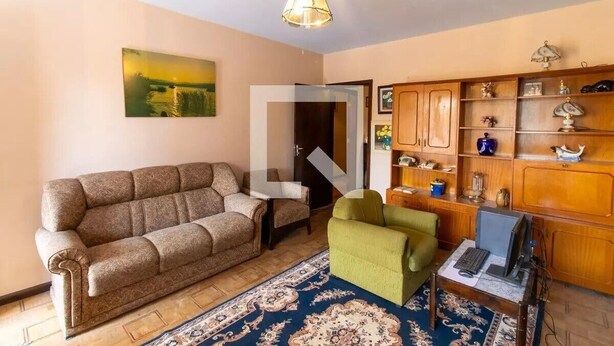 A living room with a sofa, armchair, and a small table with a laptop. There is a wooden cabinet with decorative items and a painting on the wall.