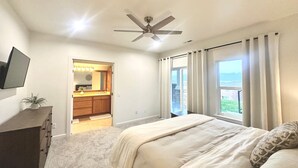 Une chambre à coucher avec un lit, un ventilateur de plafond, un téléviseur fixé au mur, une salle de bain avec un meuble-lavabo et une vue sur l’extérieur.