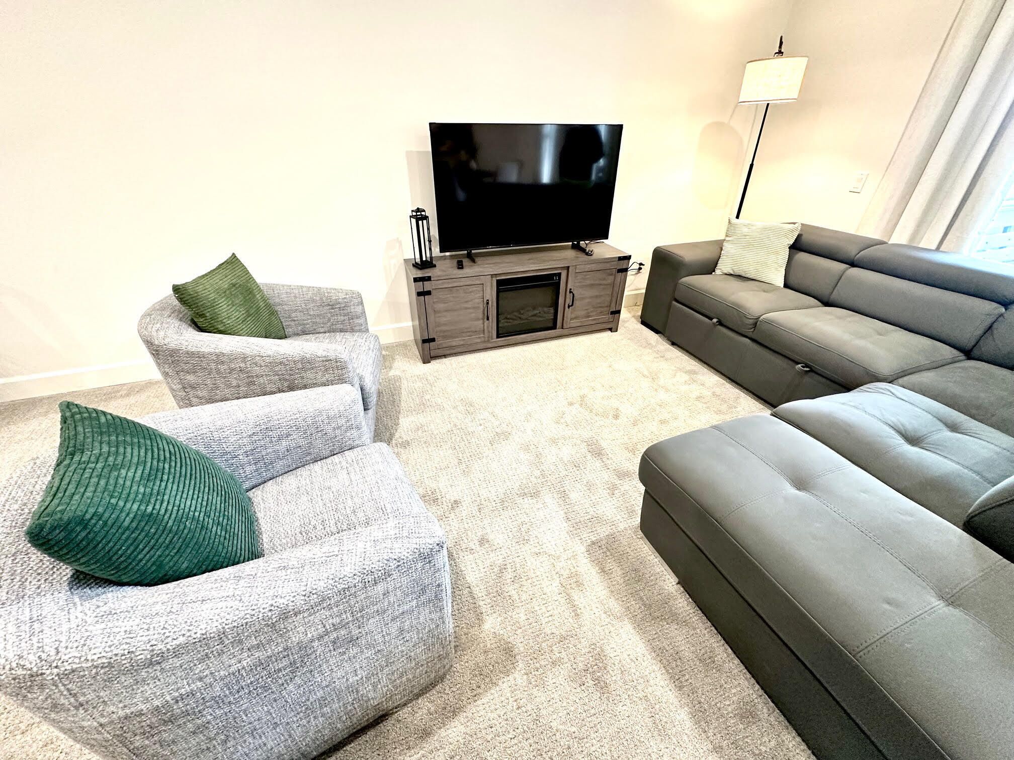 A living room with a grey sectional sofa, two grey armchairs with green cushions, a TV on a wooden stand, and a floor lamp.