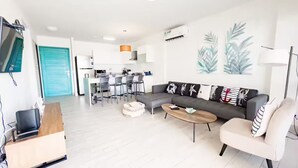 A modern living room with a grey sofa, a wooden coffee table, and a white armchair. There is a dining area with a table and chairs, and a kitchen in the background. - Sanctuary Sea View -S1-106- 2 bed, 2 bath (Punta Chame)