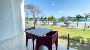 A balcony with a table and chairs, overlooking a beach with palm trees and thatched umbrellas. - Sanctuary Sea View -S1-106- 2 bed, 2 bath (Punta Chame)