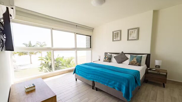 A bedroom with a bed, a bedside table, a nightstand, a TV mounted on the wall, and a window with a view of greenery. - Sanctuary Sea View -S1-106- 2 bed, 2 bath (Punta Chame)