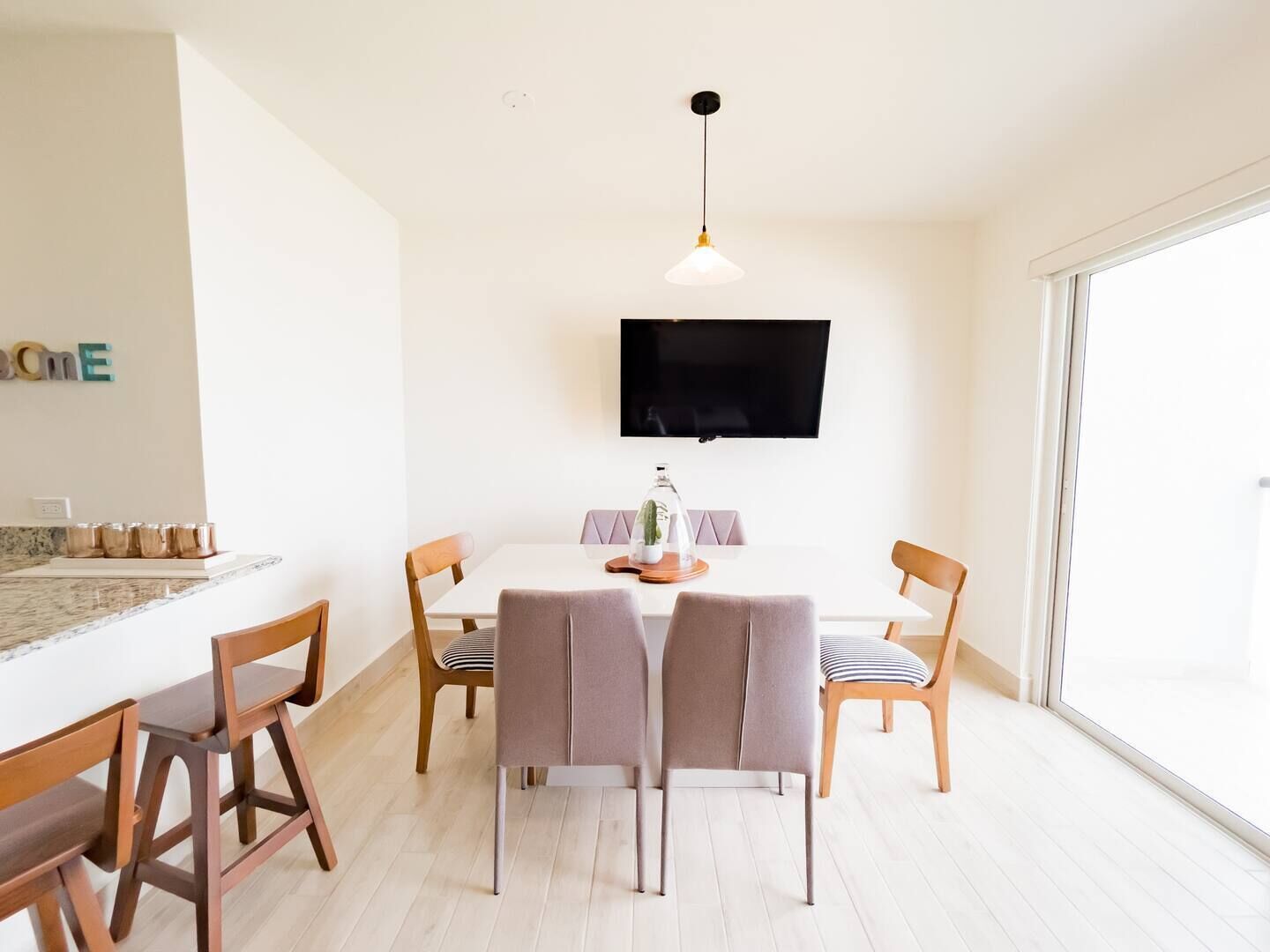 A modern dining area with a white table, four chairs, a television above, and a large window.