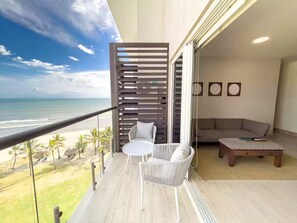 Un balcón con vista a la playa, una silla de mimbre blanca, una mesita redonda y un sofá.