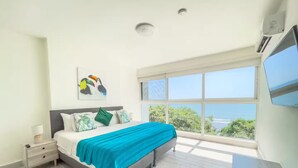 A bedroom with a large window, a bed with a blue blanket, a nightstand, and a painting on the wall. - Sanctuary Reef -S2-507- 2 bed, 2 bath (Punta Chame)