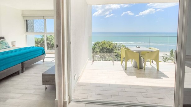 A balcony with a view of the ocean, a bed with a blue bedspread, and a table with yellow chairs. - Sanctuary Reef -S2-507- 2 bed, 2 bath (Punta Chame)