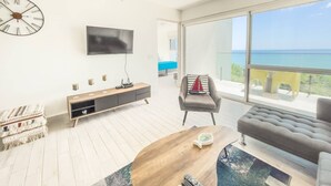 A modern living room with a wooden coffee table, a grey sofa, a TV mounted on the wall, and a large window offering a view of the sea. - Sanctuary Reef -S2-507- 2 bed, 2 bath (Punta Chame)