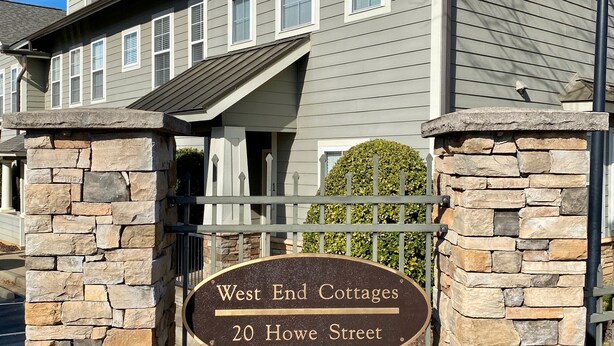 A two-story building with a stone entrance gate, a sign reading "West End Cottages 20 Howe Street," and a clear blue sky in the background. - Reedy River Retreat - Walk to Everything Downtown (Greenville)