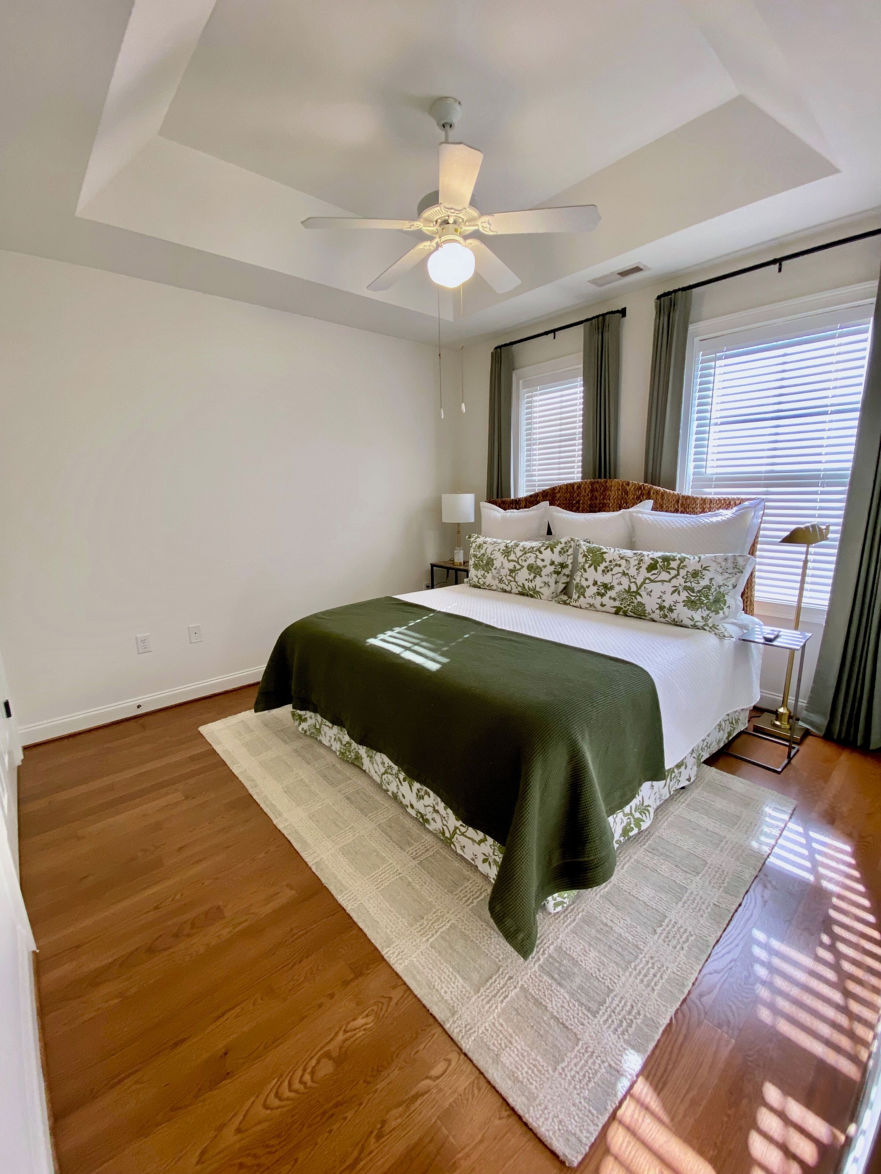 A bedroom with a bed, a ceiling fan, a window with blinds, and a nightstand with a lamp.