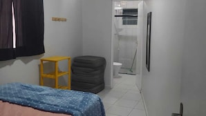 Habitació d'hotel estreta amb un llit, un tamboret petit, un gresol i una zona de dutxa.
