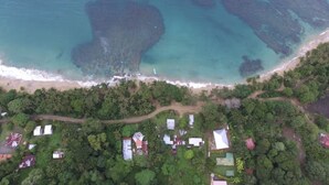 Luchtbeeld van een kustgebied met een strand, helder turquoise water en een dichtgroen bos. Er staan verschillende gebouwen met uiteenlopende dakvormen en -maten.
