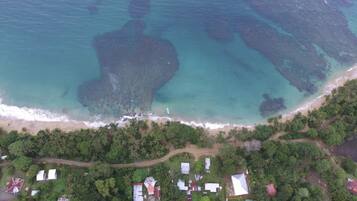 An aerial view of a coastal area with a beach, clear turquoise waters, and a dense green forest. There are several buildings with varying roof shapes and sizes.