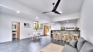 Ein geräumiger Wohnbereich mit grauem Sofa, einem Esstisch mit Stühlen, einer Küche mit Bar und einem Deckenventilator.
