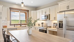 A modern kitchen with white cabinets, stainless steel appliances, a large island with a vase, and a view of the outdoors through a window. - New! Modern Retreat, Fireplace, Mountain Views, Hike, Fish, Bozeman, Yellowstone (Bozeman)