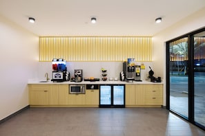 Zona de recepción de un hotel moderno con un diseño limpio y minimalista, que incluye un mostrador de recepción, una máquina de café, un refrigerador y una puerta de vidrio que da al exterior.